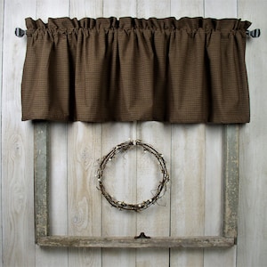 May include: Brown gingham valance with ruffles hangs on a window with a rustic wooden frame. A twig wreath with white berries hangs in the center of the window.