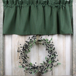 May include: A green and white striped valance with a gathered ruffle hangs above a black berry wreath on a white wood background.
