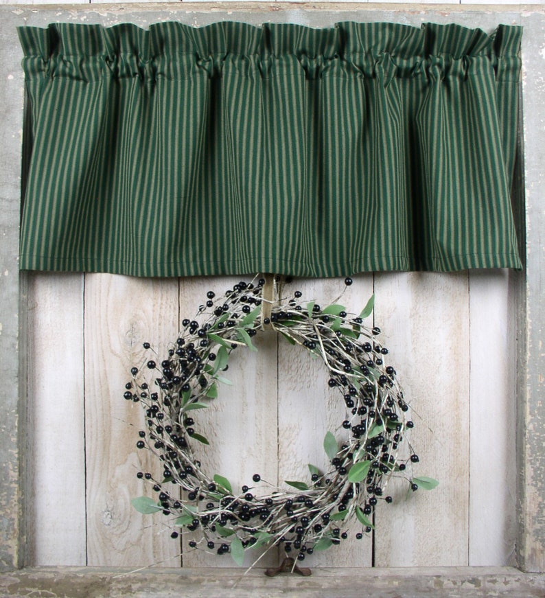 May include: A green and white striped valance with a gathered ruffle hangs above a black berry wreath on a white wood background.