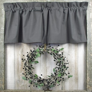 May include: A gray fabric valance with pleats hangs above a black berry wreath with green leaves. The wreath is hanging on a white wooden wall.
