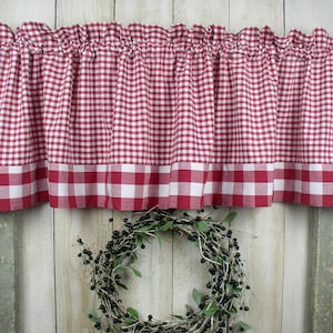 May include: Red and white gingham valance with a red trim. The valance is hung on a curtain rod and is gathered in the center. The valance is in front of a white wood background.