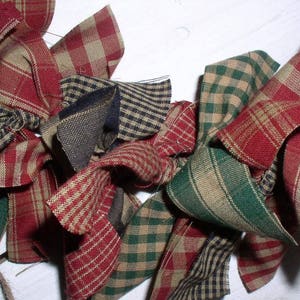 May include: Close-up of a rustic garland made of fabric bows in various patterns and colors. The bows feature red, green, tan, and navy blue plaid and striped designs. The garland is likely used for holiday or farmhouse-style decor.