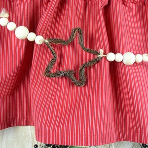 May include: A red and white striped fabric with a decorative garland. The garland features white wooden beads and a star-shaped element made of brown twigs. The fabric is gathered, creating a textured appearance.