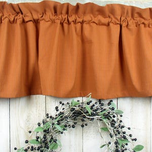 May include: A rust-colored valance with a subtle striped pattern hangs above a white wooden surface. The valance has a gathered ruffle at the top and a straight bottom edge.