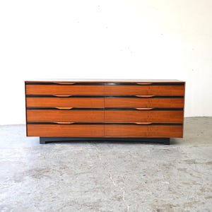 May include: A mid-century modern dresser with six drawers made of wood with a dark wood veneer. The dresser has a black base and is sitting on a concrete floor.