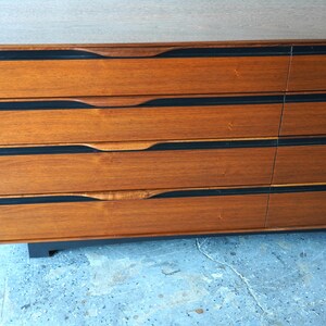 May include: A wooden dresser with six drawers. The drawers have black trim and curved handles. The dresser is sitting on a light blue surface.