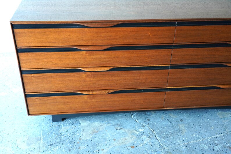 May include: A wooden dresser with six drawers. The drawers have black trim and curved handles. The dresser is sitting on a light blue surface.