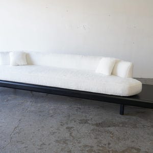 May include: A white, plush sofa with black wooden legs and a matching black wooden platform. The sofa has two white pillows on the back and one white pillow on the front.