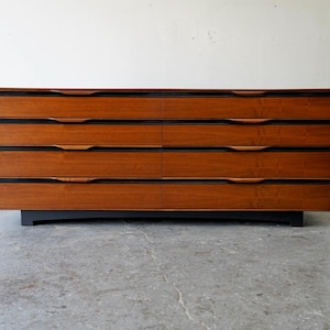 May include: A six-drawer dresser made of wood with a dark brown finish. The dresser has a black base and simple, recessed handles.