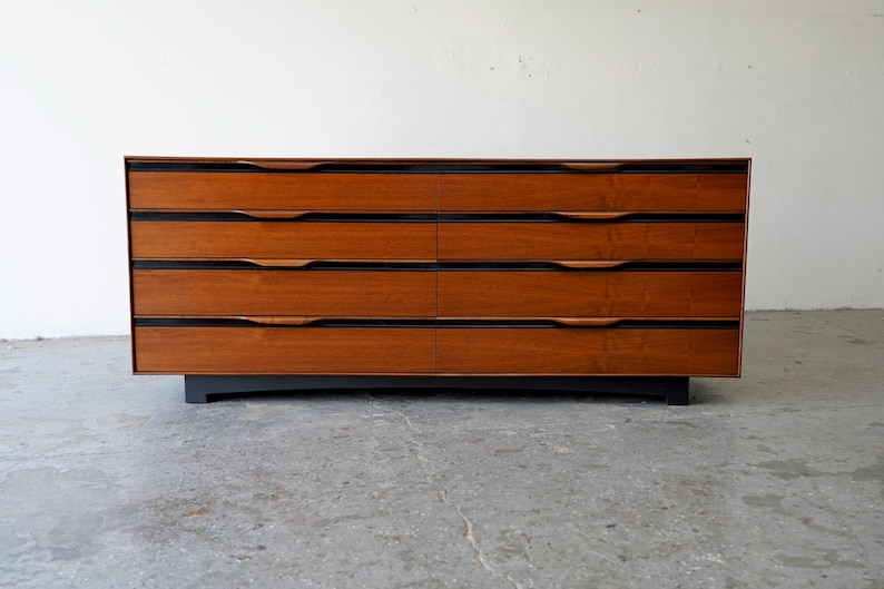 May include: A six-drawer dresser made of wood with a dark brown finish. The dresser has a black base and simple, recessed handles.