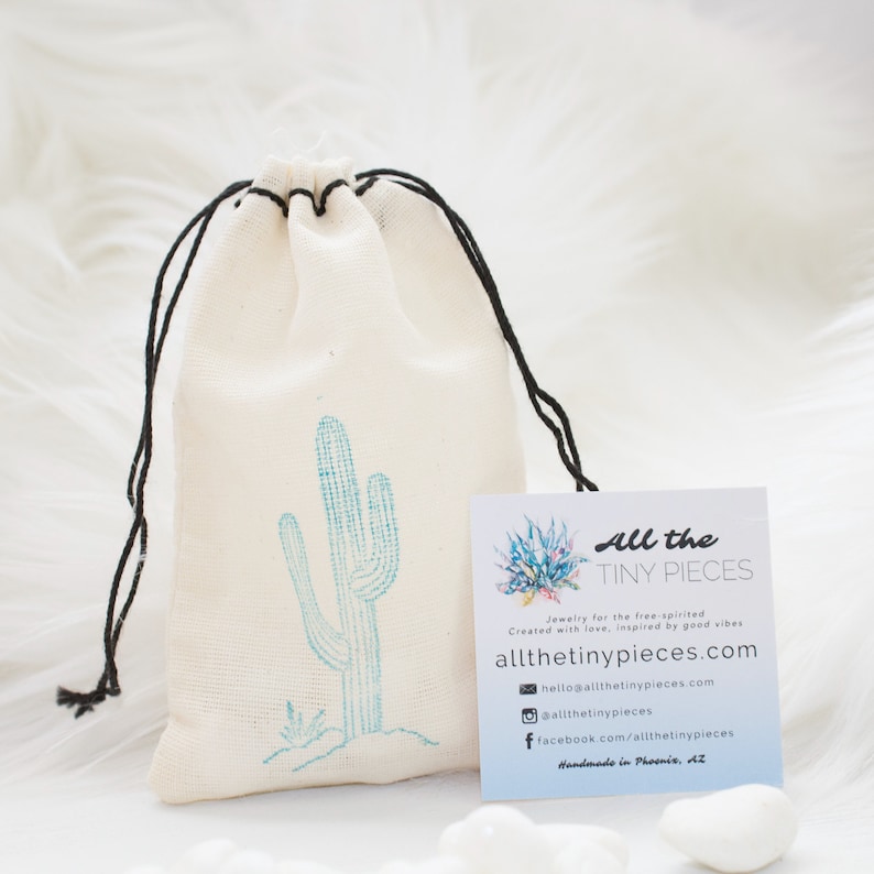 Peut inclure: Un sac en tissu cr&egrave;me avec un cordon de serrage et un motif de cactus turquoise. Une carte de visite avec le texte "All the TINY PIECES" et des coordonn&eacute;es est &agrave; c&ocirc;t&eacute;. Le sac est ferm&eacute; par un cordon noir.