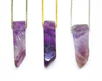 May include: Three necklaces featuring raw, elongated purple amethyst crystal pendants. The left pendant hangs on a bronze-toned chain, the center on a gold-toned chain, and the right on a silver-toned chain. Each crystal has a unique shape.