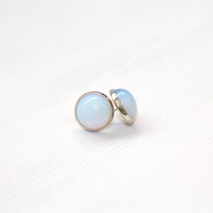 Peut inclure: Deux boucles d'oreilles rondes avec un sertissage argent&eacute; et un centre opalescent bleu clair. Les boucles d'oreilles sont pos&eacute;es sur un fond blanc, cr&eacute;ant une esth&eacute;tique &eacute;pur&eacute;e et minimaliste.