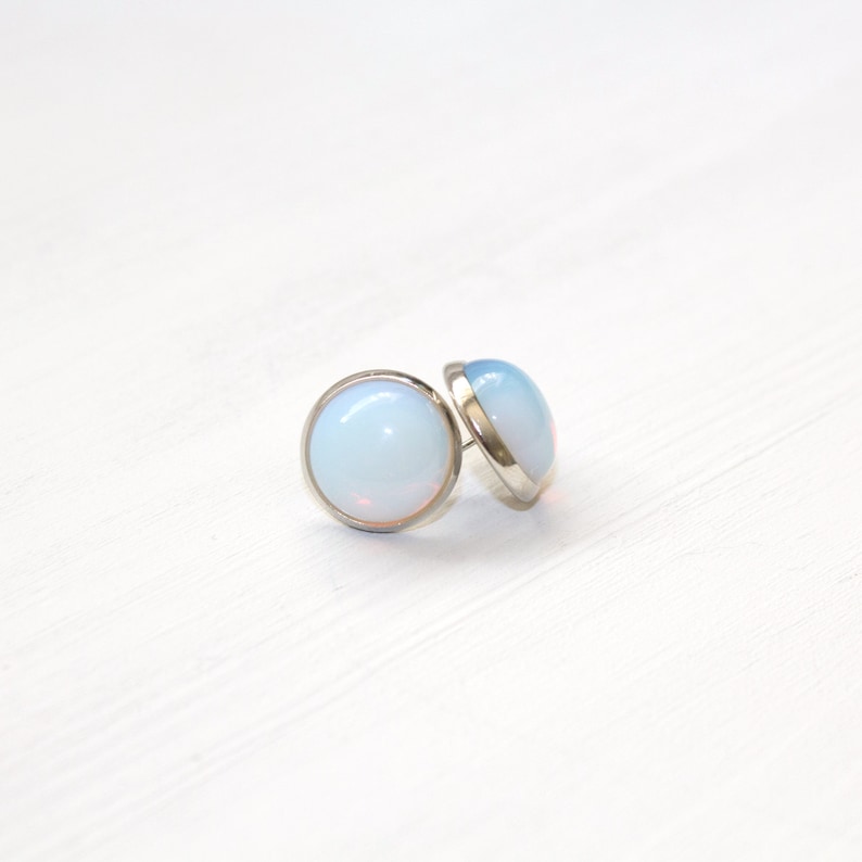 Peut inclure: Deux boucles d'oreilles rondes avec un sertissage argent&eacute; et un centre opalescent bleu clair. Les boucles d'oreilles sont pos&eacute;es sur un fond blanc, cr&eacute;ant une esth&eacute;tique &eacute;pur&eacute;e et minimaliste.
