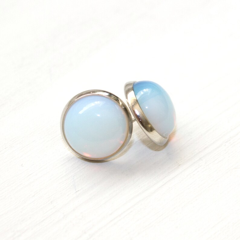 Peut inclure: Une paire de boucles d'oreilles &agrave; clous avec une pierre opalescente ronde sertie dans une lunette de couleur argent. Les pierres ont un aspect blanc laiteux doux avec de subtiles nuances de bleu et de rose. Les boucles d'oreilles sont pr&eacute;sent&eacute;es sur une surface blanche.