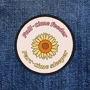 May include: Round patch with a sunflower design. The patch has a light pink background with the words "Full-time feeder" and "Part-time sleeper" in a retro font. The sunflower is yellow and orange with a brown center.