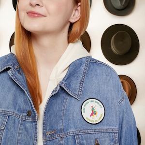 Puede incluir: Chaqueta vaquera azul con un parche redondo que dice "WHO RUNS THE WORLD? WOMEN IN LABOR". El parche presenta un gráfico colorido de dos figuras. La chaqueta se lleva sobre una sudadera con capucha blanca. Varias sombreros están montados en la pared en el fondo.