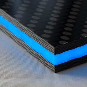 May include: A close-up of a black carbon fibre sheet with a blue LED light strip running along the edge. The sheet has a woven pattern and is illuminated by the blue light.