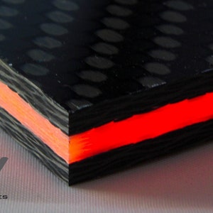 May include: A close-up of a black carbon fiber sheet with a pattern of small white circles. The sheet is layered with a red and orange translucent material, creating a glowing effect.