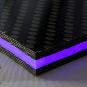 Könnte beinhalten: Nahaufnahme einer schwarzen Kohlefaserplatte mit einem violetten Leuchten, das vom Rand ausgeht. Die Platte ist geschichtet und hat eine strukturierte Oberfläche.