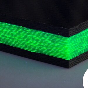 Op de afbeelding: Close-up van een zwart en groen carbonfiber blad met een gloeiende groene laag. Het blad is getextureerd en heeft een subtiele glans. De afbeelding is bijgesneden om de rand van het blad te tonen.