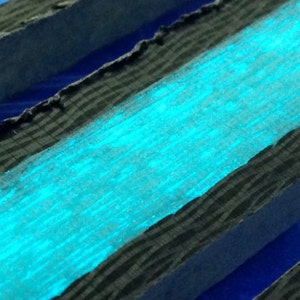 Op de afbeelding: Een close-up van een stuk hout met een laag helder turquoise blauwe verf aangebracht op het oppervlak. Het hout heeft een donkergrijze kleur en een getextureerde nerf.