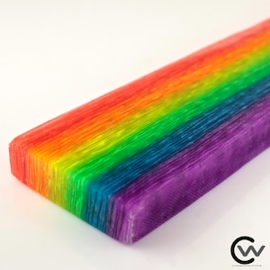 Könnte beinhalten: Ein rechteckiger Block mit einem lebendigen Regenbogen-Farbverlauf und deutlichen horizontalen Schichten in Rot, Orange, Gelb, Grün, Blau und Violett. Die Oberfläche weist über die gesamte Länge eine feine, texturierte, faserige Maserung auf.