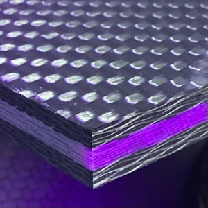 Op de afbeelding: Close-up van een gelaagd composietmateriaal van koolstofvezel. De bovenste laag heeft een geweven patroon, en de onderste laag is effen zwart. Een dunne laag paars materiaal is zichtbaar tussen de lagen.
