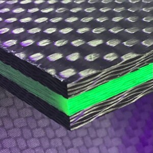 May include: Close-up of a black and green carbon fibre composite material. The material has a layered structure with a green layer in the centre.