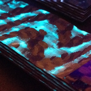 May include: A close-up of a wooden board with a blue and white glow-in-the-dark paint finish. The board is layered and has a purple glow on the bottom edge.
