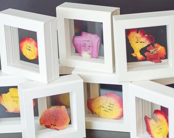 May include: A collection of white framed art pieces, each featuring a colorful flower petal with printed text. The petals display shades of red, yellow, and pink. The text includes phrases like "Listen to the call of the one."