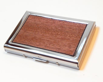 May include: A silver metal card holder with a wooden inlay. The holder has a hinged flap and a smooth, polished finish.