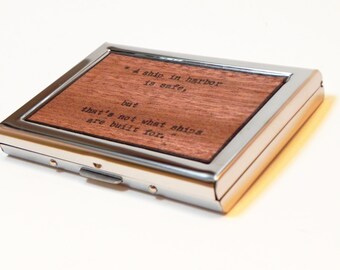 May include: A silver metal card holder with a wooden insert featuring the quote "A ship in harbor is safe, but that's not what ships are built for."