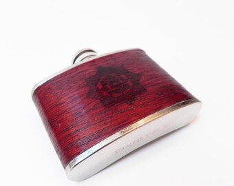 May include: A silver hip flask with a woodgrain design and a starburst pattern. The flask is labeled "Stainless Steel".