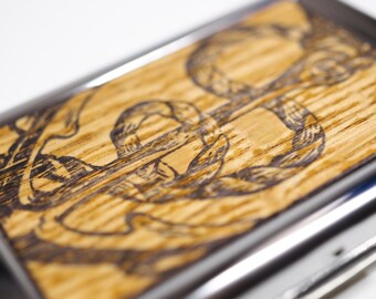 May include: A wooden cigarette case with a silver metal trim. The case has a woodgrain pattern and a design of a knot burned into the wood.