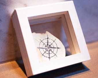 May include: A white wooden frame with a glass front displays a compass design on a piece of paper.