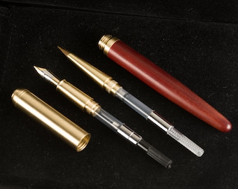 May include: Three pens on a black surface. One pen is made of wood with a red finish. The other two pens are made of metal with a gold finish. One of the metal pens has a clear plastic tube.