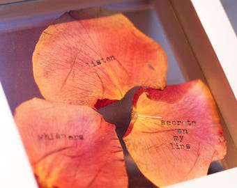 May include: Three dried rose petals in a white frame. The petals are arranged in a semi-circle. The top petal has the word "listen" written on it. The middle petal has the word "whispers" written on it. The bottom petal has the words "secrets on my lips" written on it.