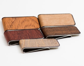May include: Five wooden money clips in different wood finishes. The clips are made of metal and have a dark brown trim. The wood finishes include light brown, dark brown, reddish brown, and a light beige.