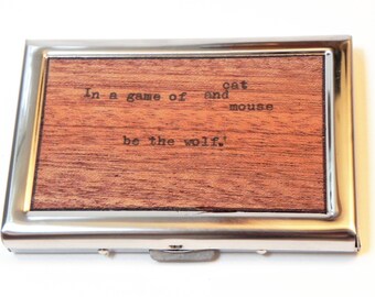 May include: A rectangular business card holder with a silver metal frame and a wooden insert. The wooden insert has the quote "In a game of cat and mouse be the wolf!" printed on it.