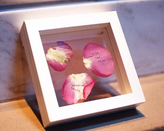 May include: A white frame with a glass front containing three dried rose petals. The petals have the following text written on them: "kiss the wind", "whisper truth", and "kiss the wind".