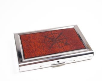 May include: A silver metal business card holder with a wood inlay featuring a compass design.