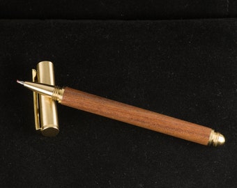 May include: A gold and wood pen with a brown wooden barrel and a gold cap and clip. The pen is lying on a black surface.