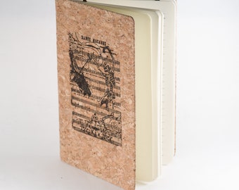 May include: A cork notebook with a black and white illustration of a skeleton playing a violin. The illustration is on a cork cover with the text "Danse Macabre".
