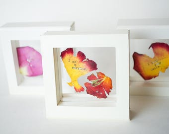 May include: Three white picture frames with dried flower petals inside. The petals are arranged in a way that creates a unique design. The frames are all the same size and shape. The text inside the frames reads "I am a warrior" and "I've got this".