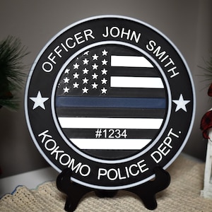 May include: A black and white round wooden sign with the American flag design, a blue line, and the text "OFFICER JOHN SMITH KOKOMO POLICE DEPT. #1234". The sign is displayed on a black stand.