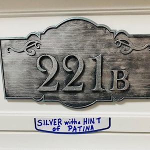 May include: Silver-colored metal address plaque with the number "221B" in bold, raised lettering. The plaque has an ornate, scrolled border and a textured surface. A handwritten note below reads "SILVER with the HINT of PATINA".