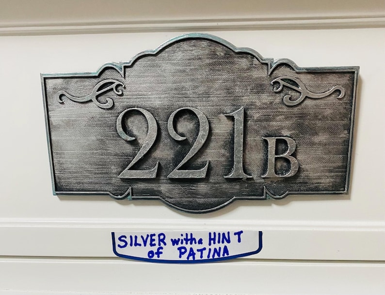 May include: Silver-colored metal address plaque with the number "221B" in bold, raised lettering. The plaque has an ornate, scrolled border and a textured surface. A handwritten note below reads "SILVER with the HINT of PATINA".