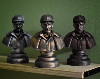 May include: Three bronze-colored busts of a man wearing a hat and coat, with a pipe in his mouth, are displayed on a wooden shelf. The busts have a detailed design and are placed against a green background.