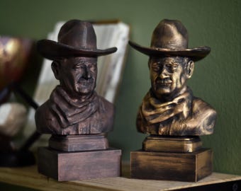May include: Two busts of a man wearing a cowboy hat and a neckerchief, each mounted on a rectangular base. The left bust has a dark bronze finish, while the right bust features a brighter, metallic gold and black finish with detailed facial features.